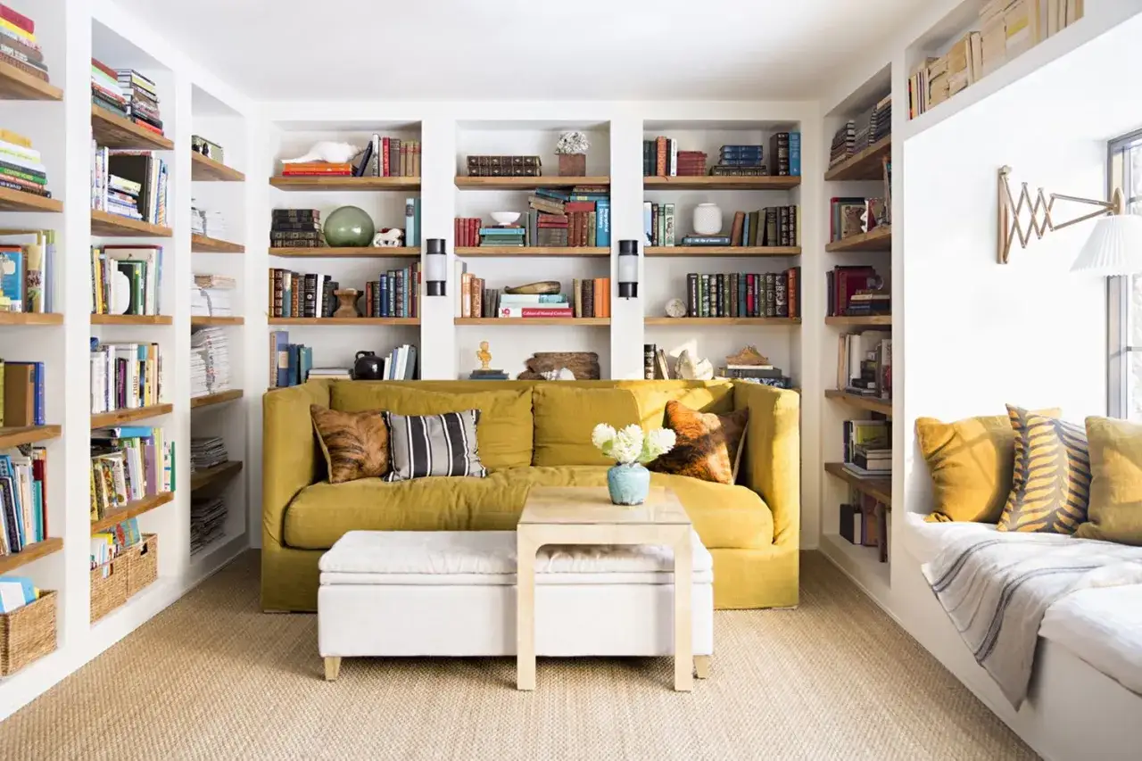 Acogedora sala de estar con sof&aacute; amarillo, mesa de centro y estanter&iacute;as llenas de libros. Un rinc&oacute;n perfecto para leer, aunque la obra menor sin licencia de las estanter&iacute;as no sea visible.