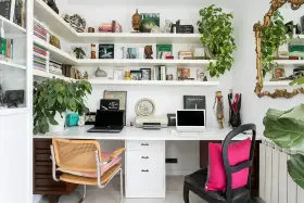 Un despacho en casa con estanterías llenas de libros y plantas. Dos portátiles, una impresora y decoración variada crean un espacio de trabajo funcional y acogedor.