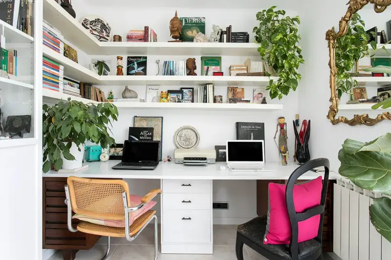 Un despacho en casa con estanterías llenas de libros y plantas. Dos portátiles, una impresora y decoración variada crean un espacio de trabajo funcional y acogedor.