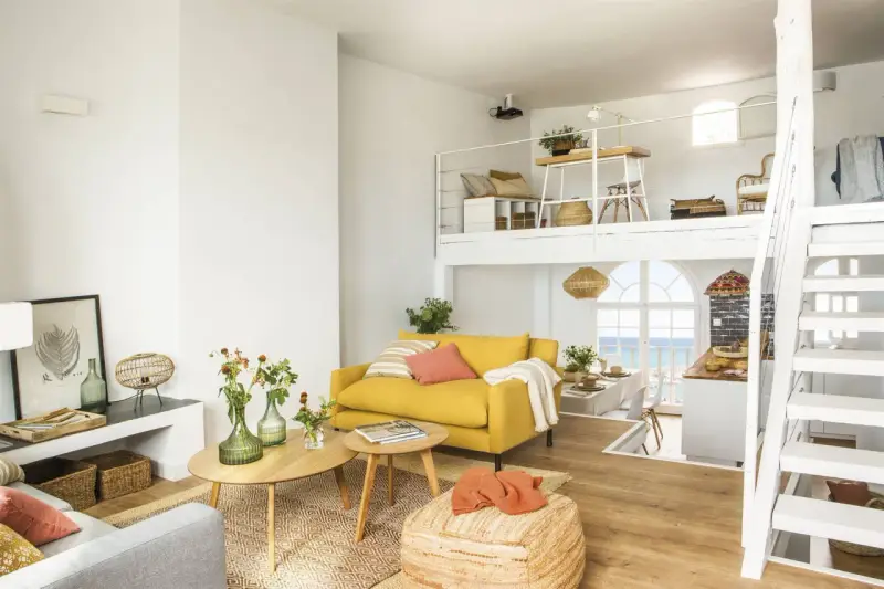 Un salón luminoso con sofá amarillo, mesas de madera y una escalera blanca que lleva a un altillo. Parece uno de esos **casas de cuento** con vistas al mar.
