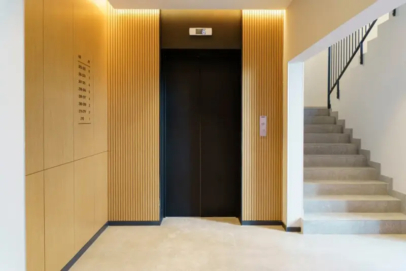 Portal moderno con ascensor negro y escaleras de mármol. Ideas para como decorar un portal de vecinos con paneles de madera y luz cálida.